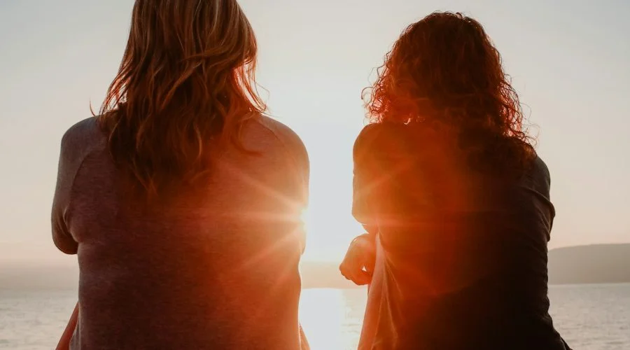 two woman sitting on beach sand while facing sunlight