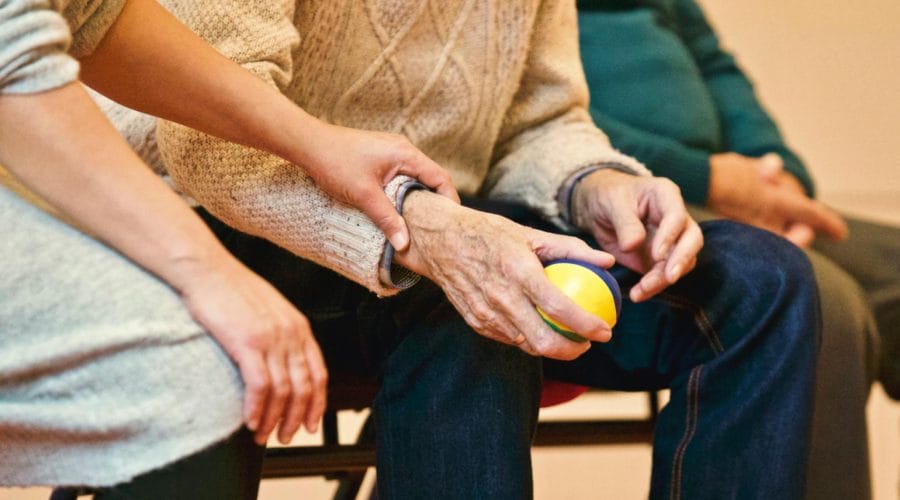 An elderly person receives support from a caregiver, holding hands indoors, showcasing compassion.