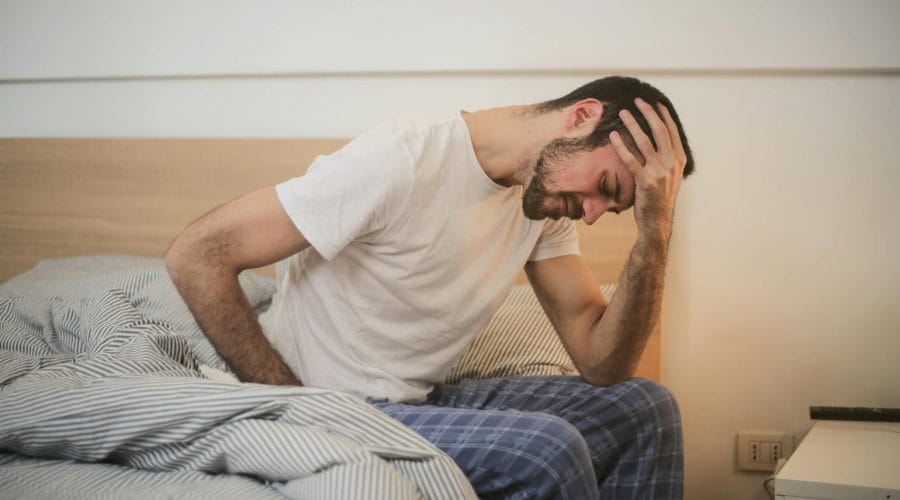 A young man in pajamas holding his head, sitting on a bed, appears to be experiencing a headache.