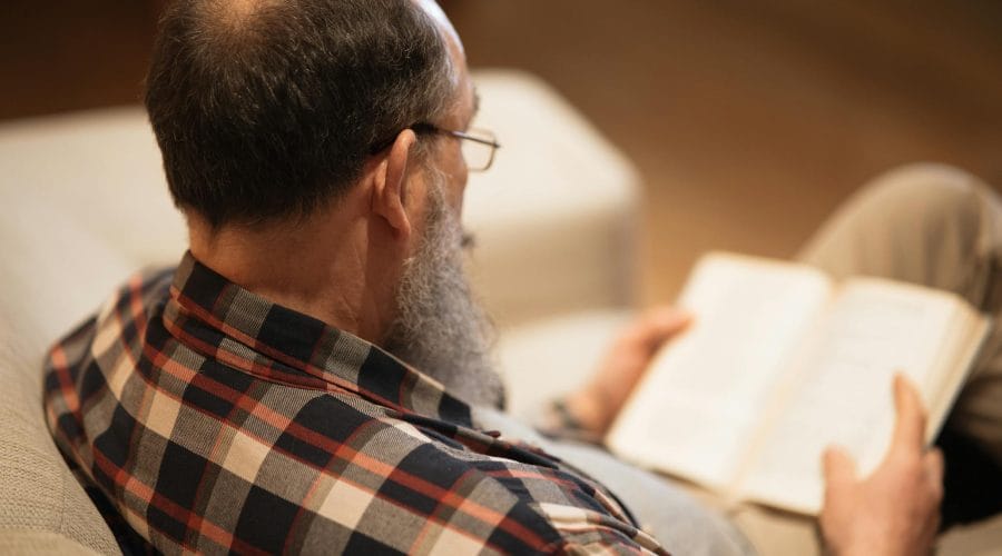 Senior man with a beard reading a book on the couch, creating a cozy atmosphere at home.