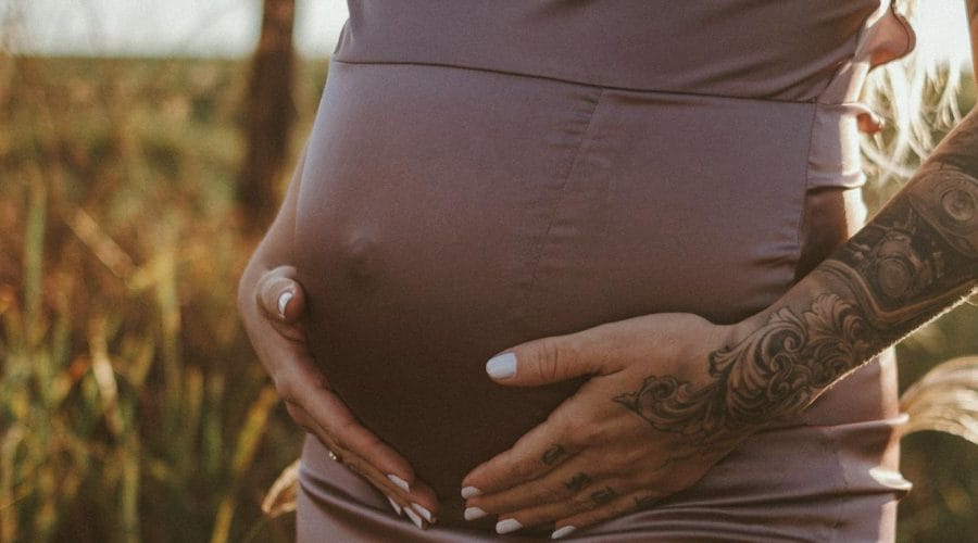 Close-up of a pregnant woman in a purple dress holding her belly in a sunlit field.