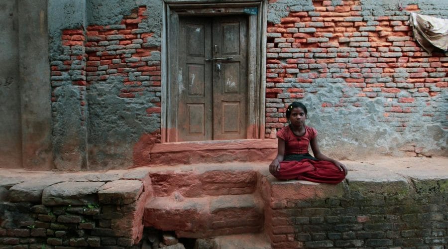 A young girl sits outside a rustic brick house in a contemplative posture.
